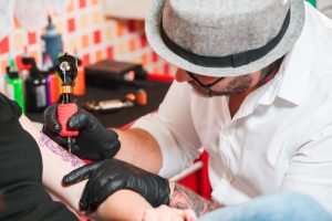 ros plan sur un tatoueur professionnel portant un chapeau et des lunettes, appliquant de l'encre violette sur l'avant-bras d'un client dans un salon de tatouage lumineux, avec des bouteilles d'encre colorées en arrière-plan