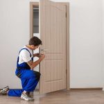 Un technicien en bleu de travail répare une serrure qui accroche sur une porte en bois.